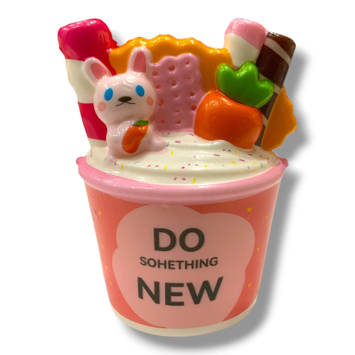 A novelty squishy toy shaped like a pink ice cream cup with the text 'DO SOMETHING NEW' on the front. The cup is topped with a white bunny holding an orange carrot, a pink cracker, a pink-and-white lollipop, an orange carrot, and a brown-and-white chocolate stick, all on a white background.