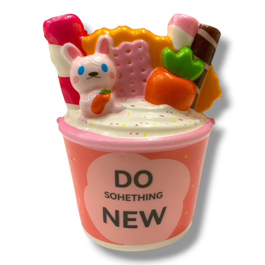 A novelty squishy toy shaped like a pink ice cream cup with the text 'DO SOMETHING NEW' on the front. The cup is topped with a white bunny holding an orange carrot, a pink cracker, a pink-and-white lollipop, an orange carrot, and a brown-and-white chocolate stick, all on a white background.